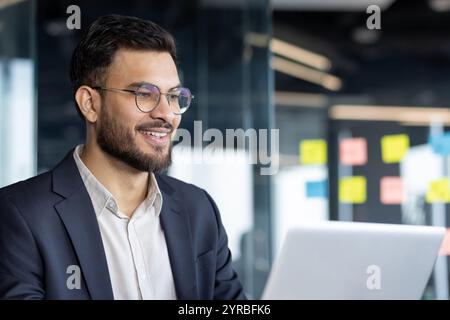 Un homme d'affaires confiant est souriant alors qu'il travaille sur son ordinateur portable dans un cadre de bureau lumineux. Son expression traduit le professionnalisme et la concentration dans un environnement d'entreprise moderne et productif. Banque D'Images