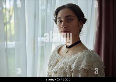 Portrait de jeune femme classique regardant la caméra avec l'expression du visage doux debout par la fenêtre tout en portant la robe délicate de dentelle, espace de copie Banque D'Images