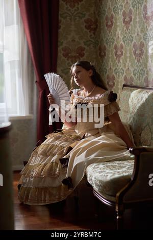 Portrait vertical pleine longueur de jeune dame élégante dans la robe bouffante classique assis sur le canapé avec ventilateur à la main contre le mur orné dans l'intérieur vintage Banque D'Images