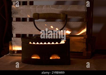 Vieux fer, chauffé par des charbons chauds. Au crépuscule, vous pouvez voir la lumière des charbons brûlants. Debout à côté d'une boîte en bois. Banque D'Images