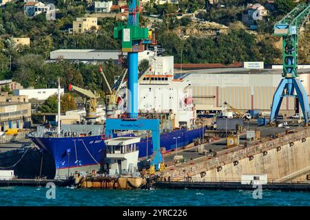 Marseille, France - 03 décembre 2024 : chantier naval avec grues imposantes et un navire amarré. Bâtiments résidentiels et collines verdoyantes en arrière-plan se mélangent Banque D'Images