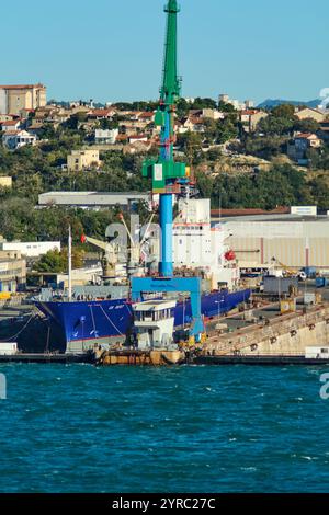 Marseille. France - 03 décembre 2024 : un chantier naval industriel avec des grues et un navire amarré, encadré par des bâtiments résidentiels et de la verdure à flanc de coteau, Banque D'Images