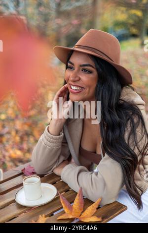 Souriante jeune femme latine portant un chapeau élégant profite d'une tasse de café à une table en bois dans un parc d'automne coloré, entouré de feuilles mortes Banque D'Images