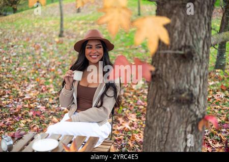 Souriante jeune femme latine portant un chapeau profite d'une tasse de café à une table en bois dans un parc d'automne coloré, entouré de feuilles tombées et d'arbres Banque D'Images