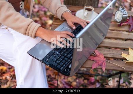 Femme latine utilisant un ordinateur portable sur une table en bois à l'extérieur en automne, avec des feuilles mortes, une tasse de café et un réveil à proximité, profitant de la saison d'automne Banque D'Images