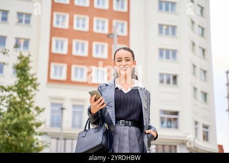 Femme souriante tenant un téléphone et un sac à main dans un cadre urbain Banque D'Images