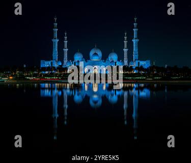 La Grande Mosquée Sheikh Zayed illuminée d'un bleu vif pendant la nuit, se reflétant sur la surface de l'eau sereine. Un chef-d'œuvre de l'architecture islamique. Banque D'Images