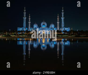 La Grande Mosquée Sheikh Zayed illuminée d'un bleu vif pendant la nuit, se reflétant sur la surface de l'eau sereine. Un chef-d'œuvre de l'architecture islamique. Banque D'Images