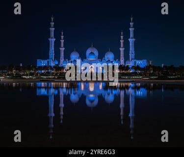 La Grande Mosquée Sheikh Zayed illuminée d'un bleu vif pendant la nuit, se reflétant sur la surface de l'eau sereine. Un chef-d'œuvre de l'architecture islamique. Banque D'Images