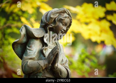 statue affectueuse et attentionnée d'une femme avec les mains priantes sur une pierre tombale d'un cimetière devant des feuilles d'automne brillantes dans un fond flou Banque D'Images