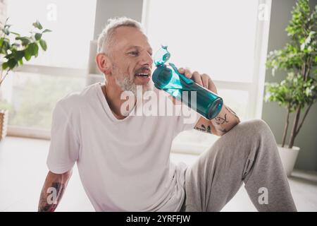 L'homme âgé aime la boisson rafraîchissante tout en se relaxant à l'intérieur dans une tenue sportive sur la journée lumineuse Banque D'Images