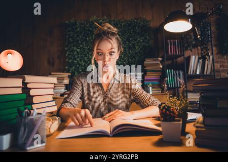 Surprise jeune femme étudiant dans un cadre de maison confortable avec des piles de livres et un éclairage chaud autour d'elle Banque D'Images