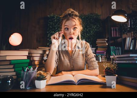 Jeune femme en chemise à carreaux surprise en étudiant à la maison tard dans la soirée, entourée de piles de livres Banque D'Images