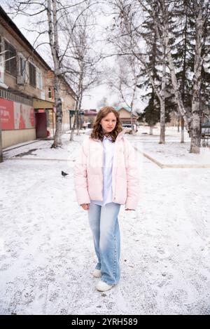 Fille russe de 15 ans en Jean bleu clair et une veste d'hiver se tient devant des bouleaux enneigés, avec une toile de fond de ville. Elle rayonne natur Banque D'Images