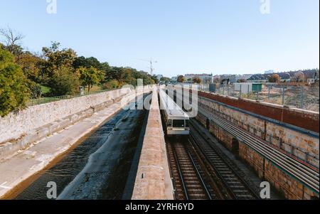 Train de métro passant entre une petite rivière et un mur de pierre à vienne par une journée ensoleillée avec des arbres et des bâtiments en arrière-plan Banque D'Images