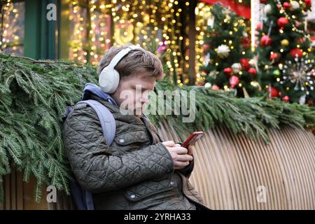 Homme dans des écouteurs assis avec smartphone sur la rue de la ville décorations de nouvel an fond, vacances d'hiver Banque D'Images