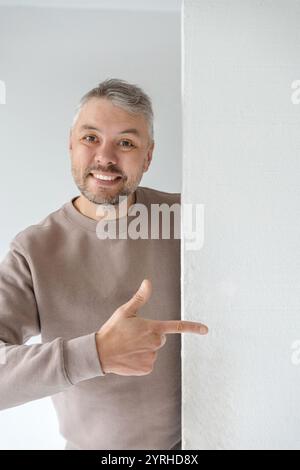 Homme sympathique dans un sweat-shirt taupe pointe vers un espace blanc vide, parfait pour ajouter du texte ou un logo. Son expression joyeuse et sa pose décontractée créent un Banque D'Images