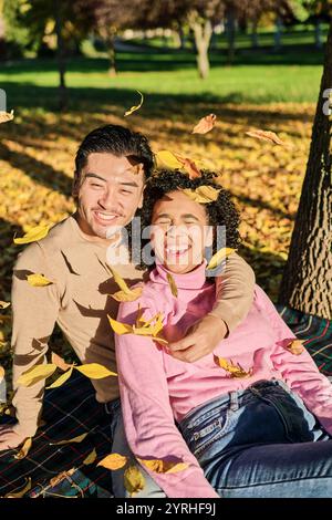 Un couple joyeux est assis ensemble sur une couverture de pique-nique, entouré de feuilles jaunes vibrantes tombant des arbres dont ils rient et embrassent la joie Banque D'Images