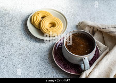 Gros plan d'une tasse à café chaude sur une assiette décorative, associée à des biscuits au beurre doré sur un petit plat, posée sur une table texturée avec une serviette en lin fo Banque D'Images