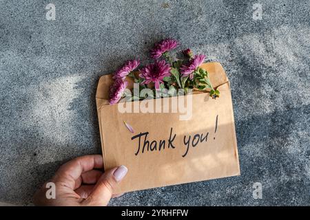 Main tenant une enveloppe avec des fleurs violettes jetant un coup d'œil et une note de remerciement manuscrite, mis sur un fond gris texturé Un symbole de gratitud Banque D'Images