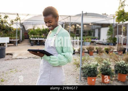 Un joyeux employé de magasin de jardin portant un tablier vérifie le stock sur une tablette numérique entourée de plantes en pot et de verdure, ils gèrent l'inventaire effique Banque D'Images