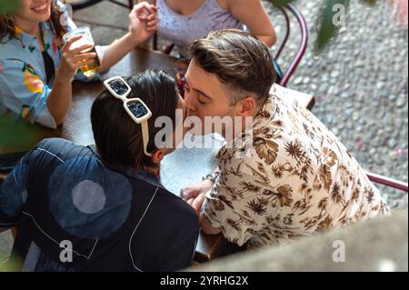Un moment intime capturé alors qu'un couple partage un baiser dans un café en plein air animé, entouré d'amis dégustant un verre par une journée ensoleillée. Banque D'Images