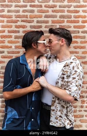 Un moment sincère entre deux hommes élégants alors qu'ils partagent un baiser et se tiennent fermement devant un mur de briques rustique. Banque D'Images