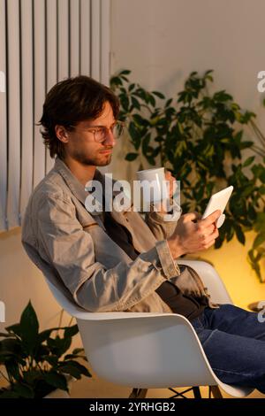 Un jeune homme en lunettes est assis dans une chaise moderne, tenant une tasse à café et une tablette entourée de plantes, il apparaît détendu dans une pièce confortable et légèrement éclairée Banque D'Images