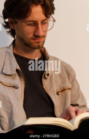 Un jeune homme avec des lunettes et une veste décontractée est absorbé dans un livre son expression focalisée suggère un engagement profond, créant une atmosphère de calme dans Banque D'Images