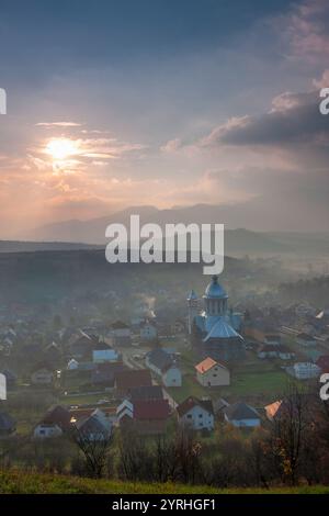 Une vue matinale sereine d'un village à Maramures, Roumanie, avec un dôme d'église sous un lever de soleil majestueux montagnes brumeuses et maisons dispersées reflètent le Banque D'Images