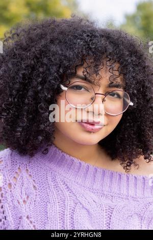 Portrait en gros plan d'une jeune femme afro-latino portant des lunettes et un pull en maille violette, souriant doucement ses cheveux bouclés encadrent son visage en exsudant Banque D'Images