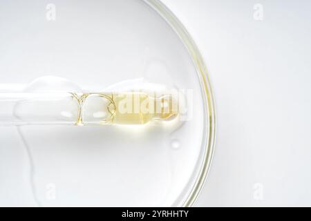 Une ampoule en verre repose sur une surface blanche, réfléchissant la lumière et contenant un liquide clair. La composition minimaliste met en valeur un esthétisme épuré et moderne Banque D'Images