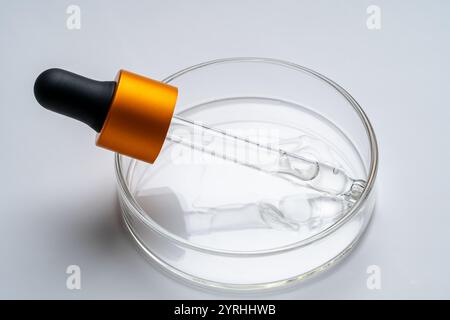 Image en gros plan d'une pipette compte-gouttes transparente avec un capuchon orange, reposant sur une boîte de Pétri contenant un liquide transparent. Idéal pour les thèmes de la science, re Banque D'Images