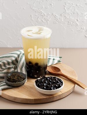 Une tasse rafraîchissante de thé moussant avec de la mousse crémeuse et des perles de tapioca sur une planche de bois accompagnée d'un bol de perles, de thé en feuilles mobiles et de bois Banque D'Images