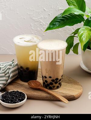 Deux délicieux thés bulles avec perles de tapioca, l'un crémeux, l'autre fruité, sur une planche de bois avec une cuillère en bois à proximité d'une plante verte ajoute une touche fraîche Banque D'Images