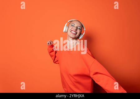 Femme adulte extatique d'âge moyen, vêtue d'orange vif, danse joyeusement tout en profitant de la musique à travers son casque sans fil blanc contre un ora Banque D'Images