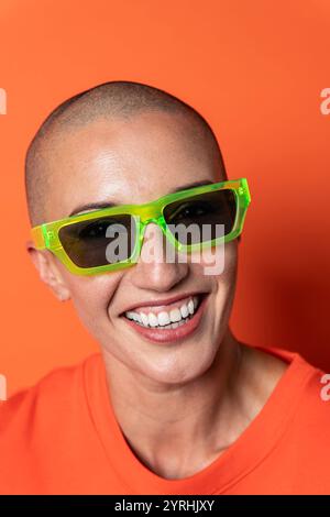 Gros plan d'une femme adulte chauve souriante portant des lunettes de soleil vert fluo sur fond orange, rayonnant de confiance et d'énergie vibrante. Banque D'Images