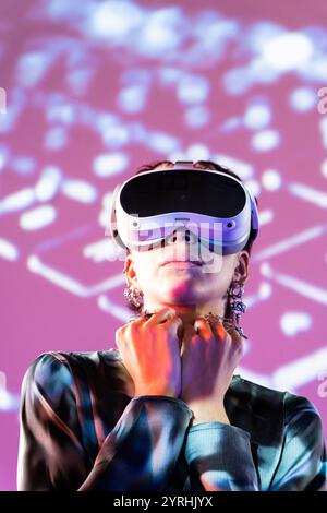 Une jeune femme dans une blouse en satin porte un casque VR blanc, debout contre un fond vibrant et inspiré numériquement qui projette des acros colorés Banque D'Images