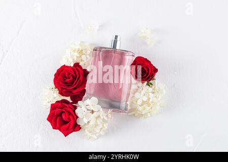 Un flacon de parfum rose est élégamment entouré de roses rouges vives et de délicates hortensias blanches, sur fond blanc immaculé, évoquant rom Banque D'Images