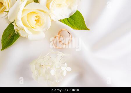 Une paire d'élégantes bagues de mariage reposant sur un fond satiné lisse, entouré de délicates fleurs blanches parfaites pour célébrer l'amour et l'engagement Banque D'Images