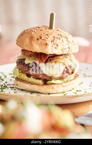 Un hamburger appétissant avec fromage fondu, bacon croustillant, laitue fraîche sur un pain aux graines de sésame, garni de ciboulette et photographié sur une table en bois Banque D'Images