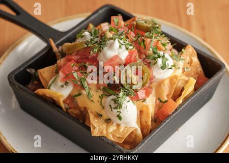 Gros plan de nachos salés recouverts de fromage fondu, de salsa fraîche, de crème sure, de jalapenos et de coriandre, servi dans une poêle noire parfaite pour l'heure de collation Banque D'Images