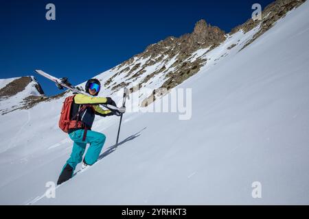 Une femme équipée de matériel de ski monte une montagne enneigée abrupte portant une tenue vibrante et portant des skis sur son dos, elle incarne l'aventure et la dissuasion Banque D'Images