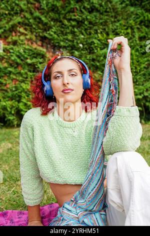 Une femme trans avec des cheveux rouges et des écouteurs bleus embrasse une écharpe colorée tout en étant assise à l'extérieur elle respire la confiance et la fierté, entourée de vert Banque D'Images