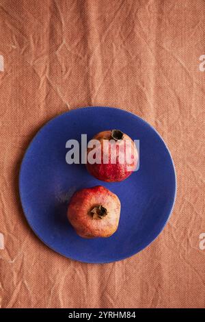 Deux grenades mûres reposent sur une plaque bleu vif placée sur un fond de tissu brun texturé. Le contraste souligne la couleur naturelle du fruit, addi Banque D'Images