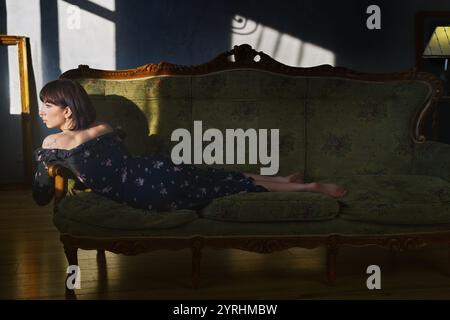 Une femme dans une robe florale se détend sur un canapé vert antique, baignée dans l'ombre dramatique et la lumière chaude du soleil. Le décor vintage évoque un sentiment de nostalgie Banque D'Images