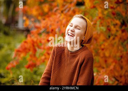 Un enfant joyeux dans un pull confortable et bonnet profite d'un rire au milieu des feuilles d'automne vibrantes dans un parc de la ville les couleurs chaudes capturent l'essence de l'automne Banque D'Images