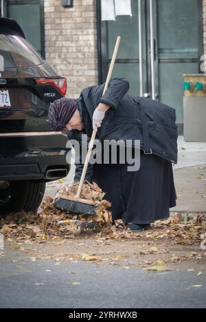 Une femme juive orthodoxe modestement vêtue dégage les feuilles du trottoir devant son entreprise. Dans une rue latérale de Brooklyn, New York. Banque D'Images