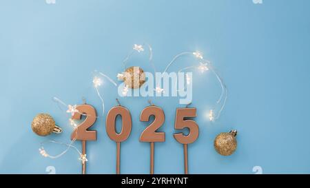 Bougies festives dorées 2025 avec des étoiles lumineuses se trouvent avec des boules dorées sur un fond bleu. Papier peint des fêtes. Nouvel an 2025 et carte de Noël Banque D'Images