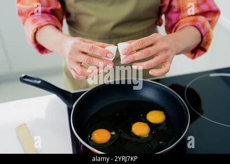 Charmante femme mature craquant des œufs dans la casserole tout en préparant le petit déjeuner dans la cuisine ensoleillée Banque D'Images